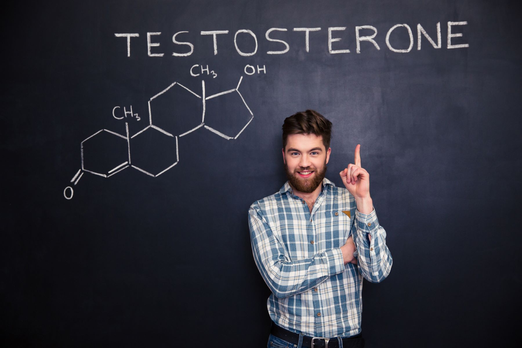 A young man standing in front of a blackboard, pointing at the word &ldquo;testosterone&rdquo; written on it above his head.